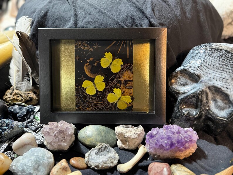 Op de afbeelding: Een ingelijst kunstwerk met een zwarte achtergrond en een gouden streep. De afbeelding toont een vrouwengezicht, een maan, sterren en gele vlinders. Het zwarte frame is omgeven door kristallen en een schedel. Een uniek decoratief stuk.