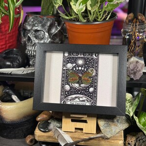 May include: A framed artwork featuring a butterfly with orange, green, and brown wings, set against a black and white tarot card design. The card is labelled "The Universe" and includes celestial imagery. The frame is black and the artwork is displayed on a wooden easel.