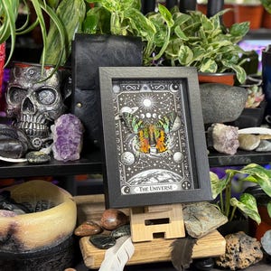May include: A framed black and white print of "The Universe" tarot card, featuring a butterfly with orange and green wings, set against a celestial background. The print is displayed on a wooden easel, surrounded by plants, crystals, and decorative items.