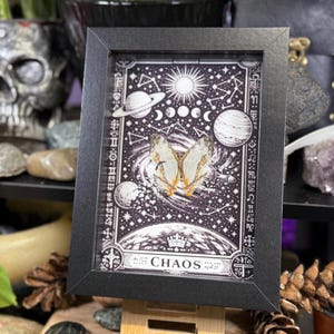 May include: A framed artwork with a black frame, depicting a black and white celestial illustration with planets, stars, and a butterfly. The word "CHAOS" is at the bottom.