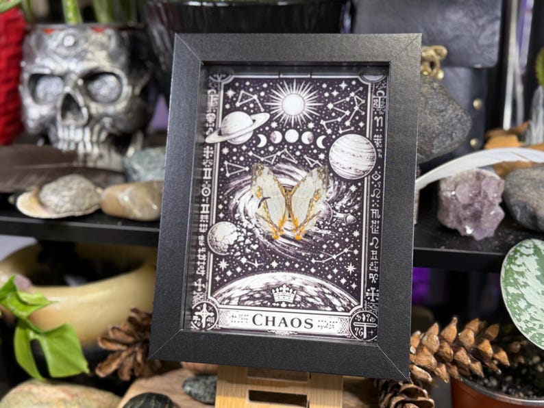 May include: A framed artwork with a black frame, depicting a black and white celestial illustration with planets, stars, and a butterfly. The word "CHAOS" is at the bottom.