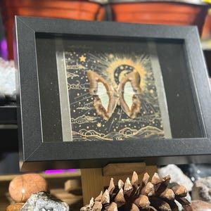 May include: A framed artwork featuring a butterfly with intricate wing patterns, set against a dark background with celestial elements. The black frame complements the artwork, creating a striking display. The artwork is displayed on a wooden stand.