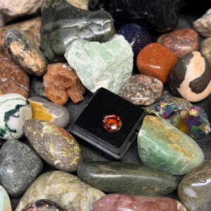 Puede incluir: Una colecci&oacute;n de piedras preciosas y rocas pulidas de varios colores, incluyendo verde, naranja y gris. Una peque&ntilde;a gema redonda de color naranja se exhibe en un estuche negro. Las piedras var&iacute;an en tama&ntilde;o y textura.