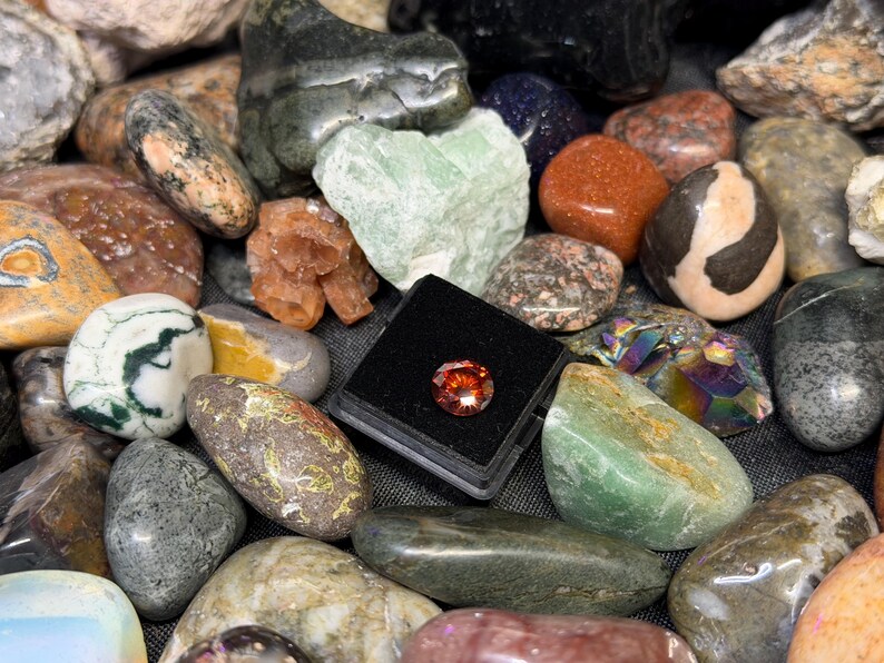Puede incluir: Una colecci&oacute;n de piedras preciosas y rocas pulidas de varios colores, incluyendo verde, naranja y gris. Una peque&ntilde;a gema redonda de color naranja se exhibe en un estuche negro. Las piedras var&iacute;an en tama&ntilde;o y textura.