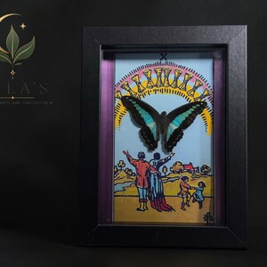Puede incluir: Un marco rectangular negro tipo caja de sombra que contiene una ilustraci&oacute;n de carta del tarot del Diez de Copas, con una mariposa azul y negra montada sobre la imagen de una familia bajo un arco&iacute;ris de diez copas doradas, con borde morado.