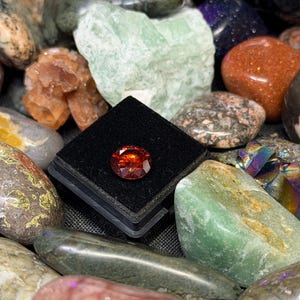 Puede incluir: Una colecci&oacute;n de piedras preciosas pulidas en varios colores y texturas. Una piedra preciosa facetada de color rojo anaranjado se exhibe en una vitrina negra. Otras piedras incluyen espec&iacute;menes verdes, marrones y multicolores.