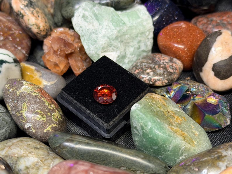 Puede incluir: Una colecci&oacute;n de piedras preciosas pulidas en varios colores y texturas. Una piedra preciosa facetada de color rojo anaranjado se exhibe en una vitrina negra. Otras piedras incluyen espec&iacute;menes verdes, marrones y multicolores.
