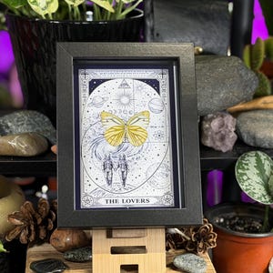 K&ouml;nnte beinhalten: Ein gerahmter Druck der Tarotkarte "Die Liebenden". Das Kunstwerk zeigt zwei Figuren, einen gelben Schmetterling und Himmelsk&ouml;rper. Der schwarze Rahmen kontrastiert mit der detaillierten Schwarz-Wei&szlig;-Illustration und erzeugt einen starken visuellen Effekt.