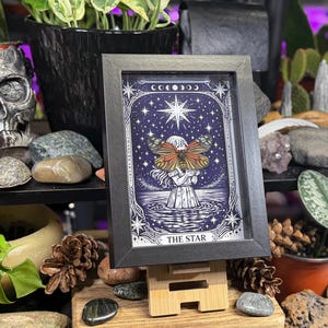 May include: A framed tarot card print of "The Star" with a black frame. The artwork depicts a figure holding a butterfly with orange and brown wings, set against a starry night sky. The print is displayed on a wooden stand.