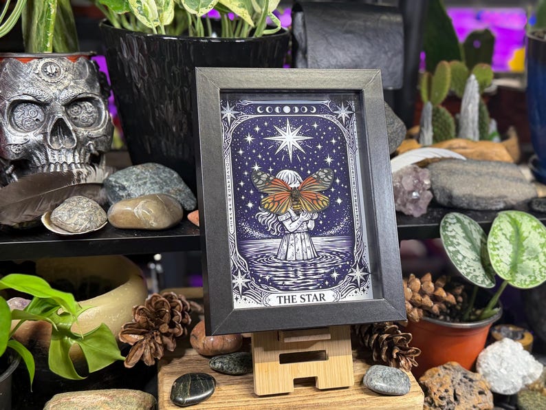 May include: A framed tarot card print of "The Star" with a black frame. The artwork depicts a figure holding a butterfly with orange and brown wings, set against a starry night sky. The print is displayed on a wooden stand.