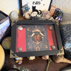 May include: A black framed display with a red background and a butterfly illustration. The frame has gold corner accents and contains several small stones. The display is surrounded by crystals, a skull, and a box with the text "Sacred Edition".