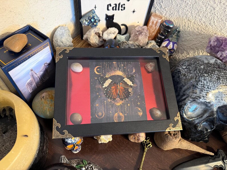 May include: A black framed display with a red background and a butterfly illustration. The frame has gold corner accents and contains several small stones. The display is surrounded by crystals, a skull, and a box with the text "Sacred Edition".