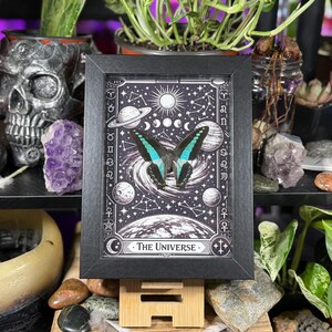 Op de afbeelding: Een zwart-wit print in een lijst met een vlinder met turquoise en zwarte vleugels, tegen een hemelse achtergrond van planeten, sterren en manen. De print is getiteld "The Universe" en wordt gepresenteerd op een houten standaard.