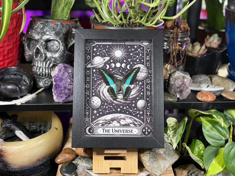 Op de afbeelding: Een zwart-wit print in een lijst met een vlinder met turquoise en zwarte vleugels, tegen een hemelse achtergrond van planeten, sterren en manen. De print is getiteld "The Universe" en wordt gepresenteerd op een houten standaard.