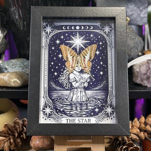 May include: Framed art print of a tarot card, "The Star." The image features a woman with butterfly wings, standing in water under a starry sky. The artwork is in a black frame.