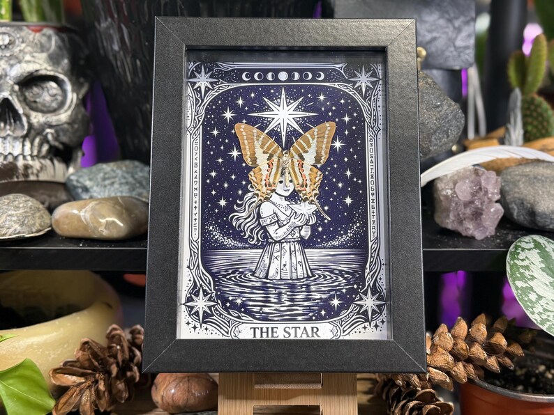 May include: Framed art print of a tarot card, "The Star." The image features a woman with butterfly wings, standing in water under a starry sky. The artwork is in a black frame.