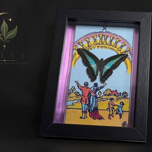 Puede incluir: Una obra de arte enmarcada con una ilustraci&oacute;n de la carta del tarot del Diez de Copas, que muestra una gran mariposa negra y azul sobre un grupo familiar, con un arco&iacute;ris de diez copas doradas encima, todo en un marco negro.