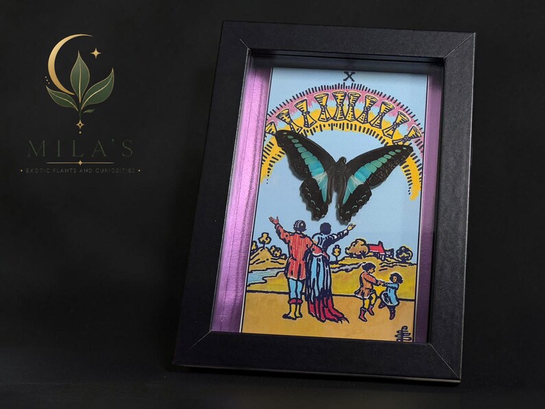 Puede incluir: Una obra de arte enmarcada con una ilustraci&oacute;n de la carta del tarot del Diez de Copas, que muestra una gran mariposa negra y azul sobre un grupo familiar, con un arco&iacute;ris de diez copas doradas encima, todo en un marco negro.