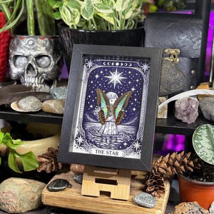 Op de afbeelding: Een ingelijste kunstdruk van de tarotkaart "The Star", met een hemelse sc&egrave;ne met een gevleugelde figuur en een grote ster. Het kunstwerk staat tegen een donkerblauwe achtergrond met sterren en manen. De lijst is zwart en de print wordt op een kleine houten ezel getoond.