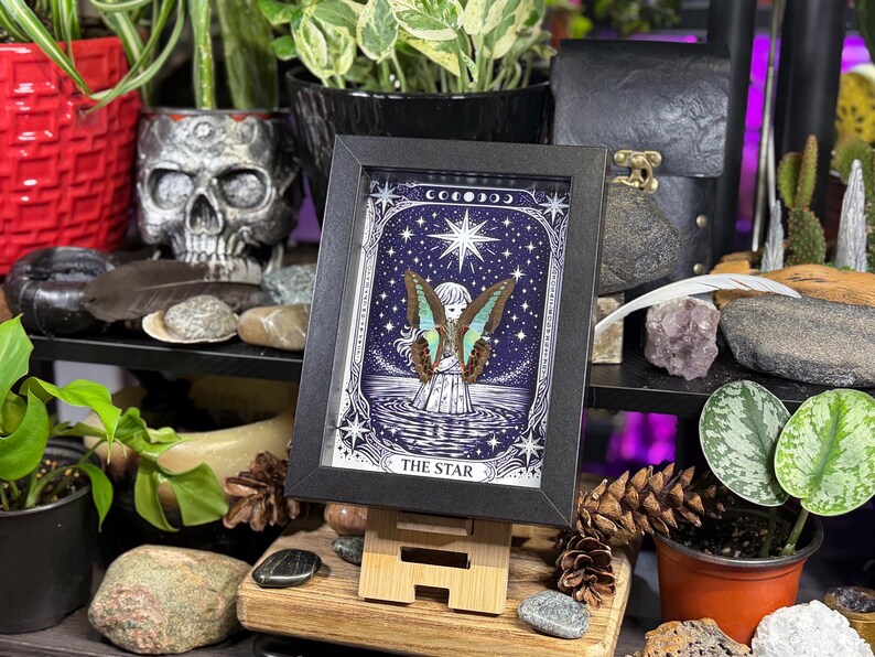 Op de afbeelding: Een ingelijste kunstdruk van de tarotkaart "The Star", met een hemelse sc&egrave;ne met een gevleugelde figuur en een grote ster. Het kunstwerk staat tegen een donkerblauwe achtergrond met sterren en manen. De lijst is zwart en de print wordt op een kleine houten ezel getoond.