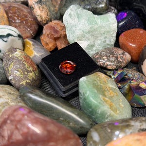 Puede incluir: Una colecci&oacute;n de piedras preciosas y rocas pulidas de varios colores y texturas. Una peque&ntilde;a gema redonda de color naranja se exhibe en una caja negra. El surtido incluye piedras verdes, marrones y moteadas.