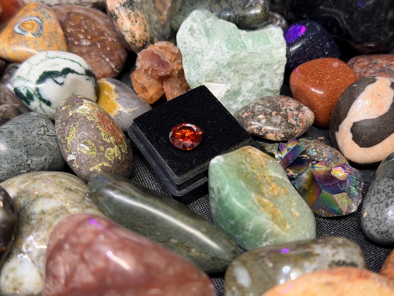 Puede incluir: Una colecci&oacute;n de piedras preciosas y rocas pulidas de varios colores y texturas. Una peque&ntilde;a gema redonda de color naranja se exhibe en una caja negra. El surtido incluye piedras verdes, marrones y moteadas.