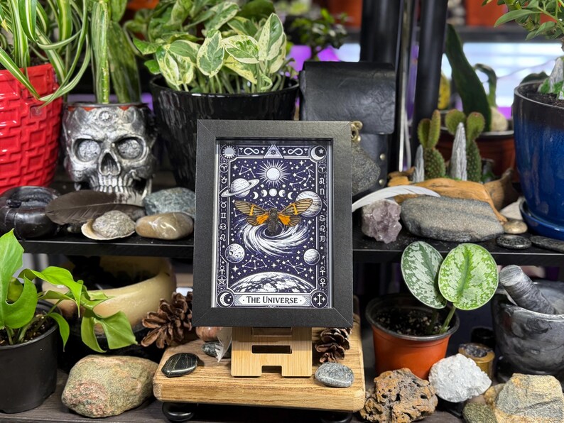 K&ouml;nnte beinhalten: Ein gerahmter Druck der Tarotkarte "Das Universum", mit einem Himmelsdesign mit Planeten, Monden und einer gefl&uuml;gelten Figur. Das Kunstwerk ist in Blau-, Wei&szlig;- und Goldt&ouml;nen gehalten und wird auf einer Holzstaffelei pr&auml;sentiert, umgeben von Pflanzen und Dekorationsartikeln.