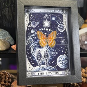 May include: Framed art print with a cosmic theme, showcasing a butterfly and astronauts. The image has a dark blue backdrop with planets and stars, and the words "THE LOVERS". The frame is dark grey.