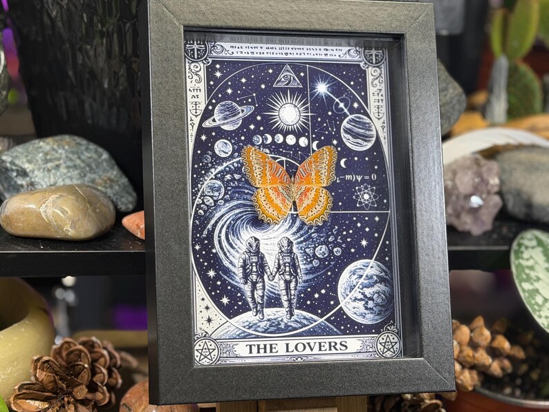 May include: Framed art print with a cosmic theme, showcasing a butterfly and astronauts. The image has a dark blue backdrop with planets and stars, and the words "THE LOVERS". The frame is dark grey.