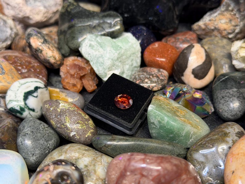 Puede incluir: Una colecci&oacute;n de varias piedras preciosas y rocas pulidas. Una peque&ntilde;a gema facetada de color rojo anaranjado se exhibe en una caja negra. Las piedras circundantes incluyen rocas verdes, marrones y blancas, con patrones y colores &uacute;nicos.