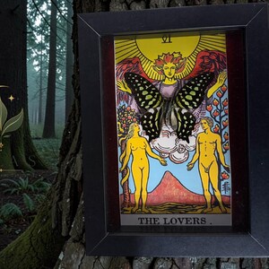 May include: A framed tarot card print titled The Lovers, featuring a central winged figure with a large butterfly overlay, flanked by two yellow human figures standing before a mountain landscape under a bright yellow sun and Roman numeral VI.