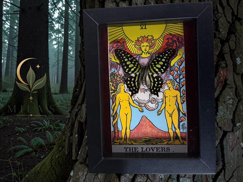 May include: A framed tarot card print titled The Lovers, featuring a central winged figure with a large butterfly overlay, flanked by two yellow human figures standing before a mountain landscape under a bright yellow sun and Roman numeral VI.