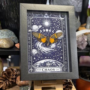 May include: Framed artwork featuring a celestial design with a butterfly and the word "CHAOS". The artwork has a dark blue background with planets, stars, and a sun. The butterfly has orange and black wings.