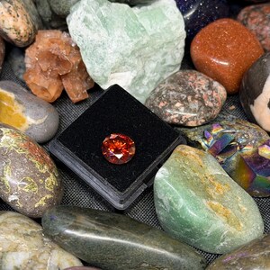 Puede incluir: Una colecci&oacute;n de diversas piedras preciosas y minerales pulidos. Una gema naranja, redonda y facetada se muestra en un estuche negro. Otras piedras incluyen variedades verdes, marrones y naranjas, con diversas texturas y patrones.