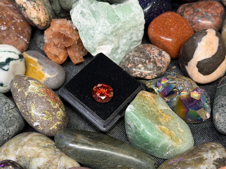 Puede incluir: Una colecci&oacute;n de diversas piedras preciosas y minerales pulidos. Una gema naranja, redonda y facetada se muestra en un estuche negro. Otras piedras incluyen variedades verdes, marrones y naranjas, con diversas texturas y patrones.