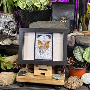 Pode incluir: Uma carta de tar&ocirc; emoldurada com uma borboleta com asas laranja e marrom, intitulada "The Lovers". A moldura preta est&aacute; em um cavalete de madeira. O fundo inclui plantas, pedras e um recipiente em forma de caveira.