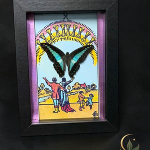 Puede incluir: Un marco negro tipo caja que contiene una ilustraci&oacute;n de carta de tarot del Diez de Copas, con una mariposa oscura central con marcas azul brillante, un arco&iacute;ris de diez copas doradas y una familia en un paisaje bajo un cielo azul.