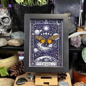 May include: A framed art print featuring a celestial design with a butterfly. The artwork is set against a dark blue background with stars, planets, and a sun. The butterfly has orange and black wings. The word "CHAOS" is at the bottom.