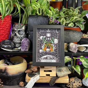 May include: A framed print with a black border featuring a celestial design with a butterfly and the word "CHAOS". The image is displayed on a wooden stand, surrounded by plants, crystals, and a skull.