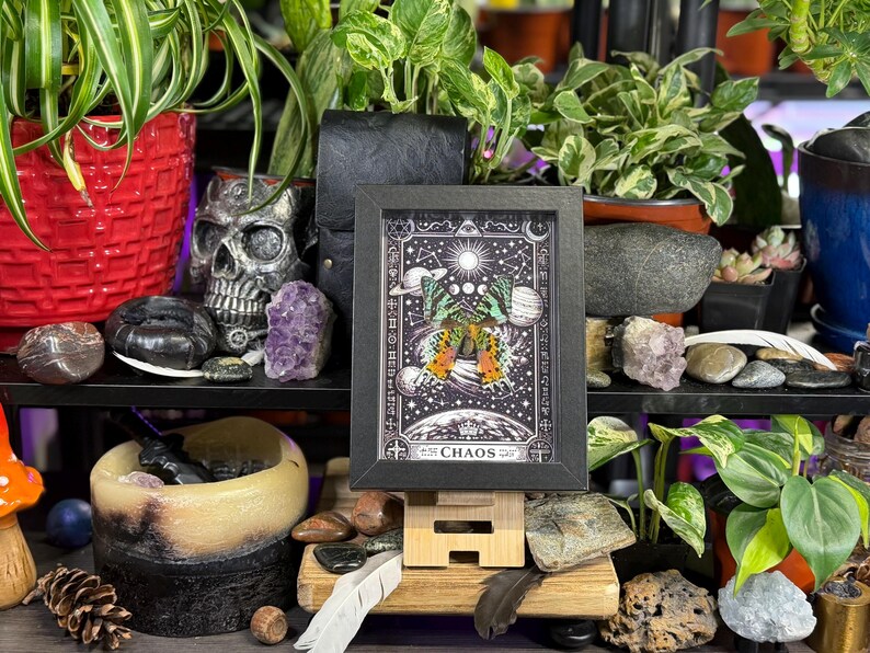 May include: A framed print with a black border featuring a celestial design with a butterfly and the word "CHAOS". The image is displayed on a wooden stand, surrounded by plants, crystals, and a skull.