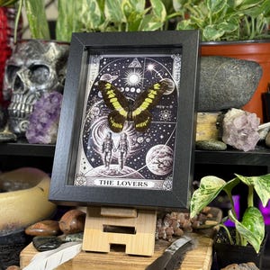 Puede incluir: Una impresi&oacute;n enmarcada que presenta una mariposa con alas amarillas y negras, sobre un fondo celestial con planetas y estrellas. La impresi&oacute;n est&aacute; etiquetada como "THE LOVERS" y se muestra en un caballete de madera.