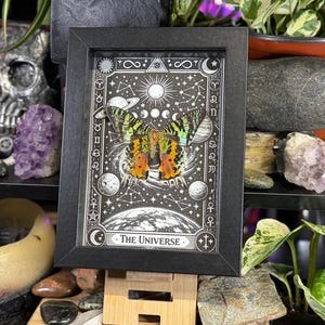 May include: Black framed art print featuring a butterfly with orange and green wings, set against a celestial background with planets, stars, and the text "The Universe". The artwork is in black and white with intricate details.