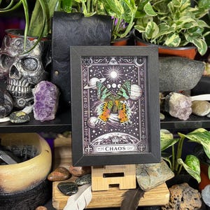 May include: A black framed artwork with a butterfly design and celestial elements, including the sun, moon phases, and planets. The artwork is set against a dark background with the word "CHAOS" at the bottom.