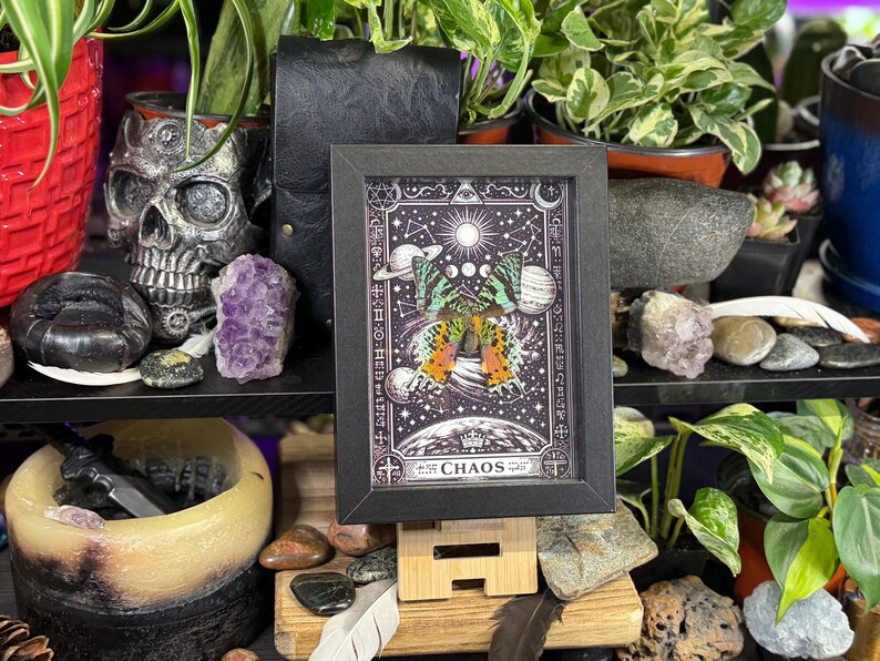 May include: A black framed artwork with a butterfly design and celestial elements, including the sun, moon phases, and planets. The artwork is set against a dark background with the word "CHAOS" at the bottom.