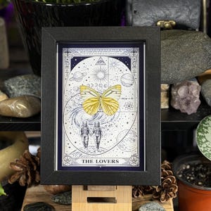 K&ouml;nnte beinhalten: Ein gerahmter Druck der Tarotkarte "Die Liebenden". Das Kunstwerk zeigt einen gelben Schmetterling, zwei H&auml;nde haltende Figuren und himmlische Elemente. Der Rahmen ist schwarz und der Druck hat einen blauen Rand mit wei&szlig;en Illustrationen. Der Hintergrund umfasst Pflanzen und Steine.