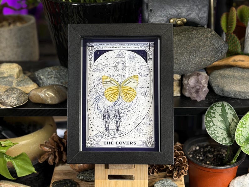 K&ouml;nnte beinhalten: Ein gerahmter Druck der Tarotkarte "Die Liebenden". Das Kunstwerk zeigt einen gelben Schmetterling, zwei H&auml;nde haltende Figuren und himmlische Elemente. Der Rahmen ist schwarz und der Druck hat einen blauen Rand mit wei&szlig;en Illustrationen. Der Hintergrund umfasst Pflanzen und Steine.