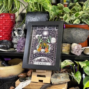 May include: A black framed artwork depicting a butterfly with orange and yellow wings, set against a celestial background. The artwork is displayed on a wooden stand, surrounded by plants and decorative items, including a skull and crystals.