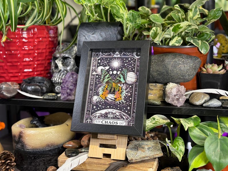 May include: A black framed artwork depicting a butterfly with orange and yellow wings, set against a celestial background. The artwork is displayed on a wooden stand, surrounded by plants and decorative items, including a skull and crystals.