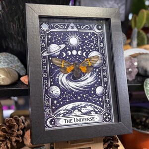 K&ouml;nnte beinhalten: Gerahmtes Kunstwerk mit einem kosmischen Design und einer Illustration einer Motte. Der schwarze Rahmen umgibt einen dunkelblauen Hintergrund mit Planeten, Monden und Sternen. Der Text "The Universe" befindet sich unten.