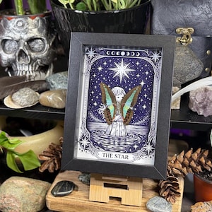 Op de afbeelding: Een ingelijste print van de tarotkaart "De Ster". Het kunstwerk toont een figuur met vlindervleugels die in het water staat onder een sterrenhemel. De zwarte lijst bevat de print, die op een houten standaard wordt getoond. Het thema is mystiek en hemels.